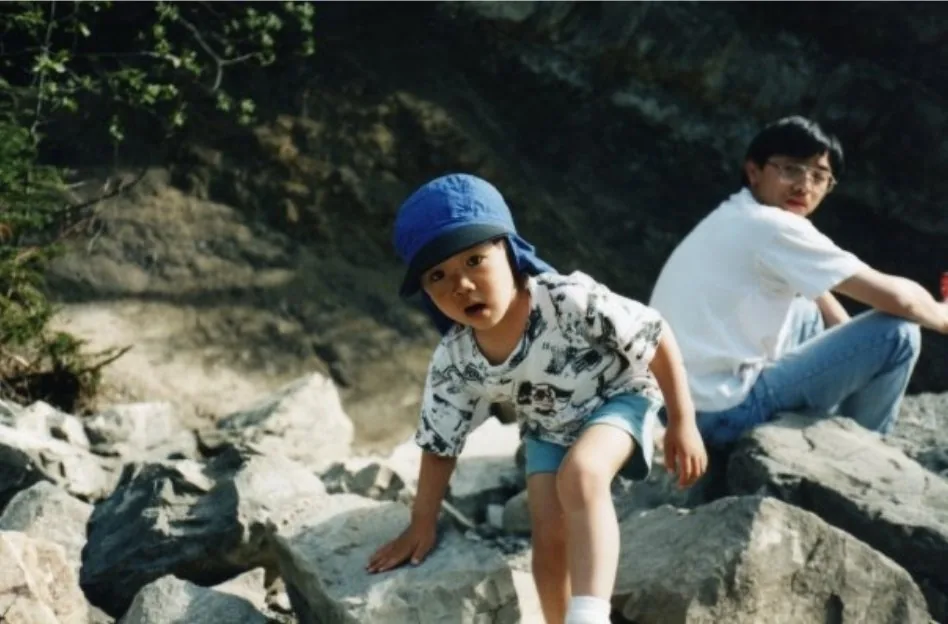 Young Brennan in a blue sun hat climbing over rocks, staring down the camera, with a man sitting on the rocks behind him.