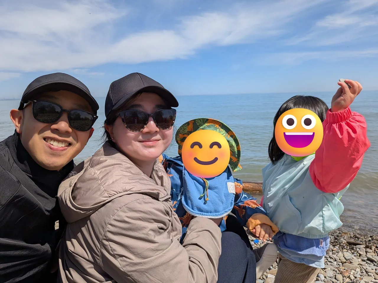 Brennan, his wife, and their two kids smiling on a pebble beach by the water on a sunny day. The kids’ faces are covered with smiley emoji.