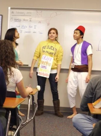 Teenage Brennan dressed as Aladdin, in a red fez, purple vest, and white pants, barefoot at the front of a classroom beside a classmate in a yellow hoodie, presenting to seated students.